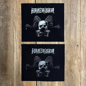 Puede incluir: Dos parches negros con la palabra "HANZAISHA" en fuente gótica. Cada parche presenta una calavera con máscara de gas, con cuernos y un fondo oscuro y texturizado. Los parches son rectangulares y tienen apariencia tejida.