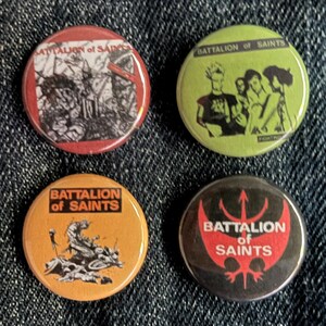 Może przedstawiać: Cztery okrągłe przypinki z napisem "Battalion of Saints" i różnymi grafikami. Przypinki są czerwone, zielone, pomarańczowe i czarne. Przypinki są na dżinsowym tle.