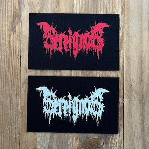 May include: Two black rectangular patches with the word "SERIGIOS" in a stylized, dripping font. One patch features red text, while the other has white text. The patches are displayed on a wooden surface.
