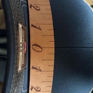 Könnte beinhalten: Eine marineblaue Baseballkappe mit einem braunen Leder-Messband, das über die Oberseite gelegt ist. Das Band zeigt Messungen in Zoll, mit den Zahlen 0 bis 3 sichtbar. Die Kappe hat ein Richardson 112-Etikett.