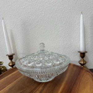 May include: A clear, lidded glass candy dish with a textured, patterned design. The dish sits on a wooden tray, flanked by two white candles in brass holders. The glass has a decorative, vintage aesthetic.
