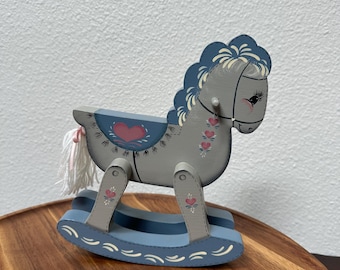 May include: A gray and blue wooden rocking horse with pink heart accents. The horse has a white mane and tail, and is painted with a folk art style. The rocking horse is a classic toy.