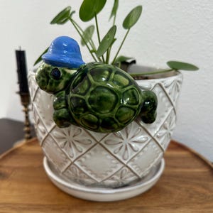 May include: A white ceramic planter with a diamond-patterned embossed texture and a matching saucer. A small, glossy green ceramic turtle figurine wearing a bright blue hat is attached to the rim of the planter, facing toward the left side.