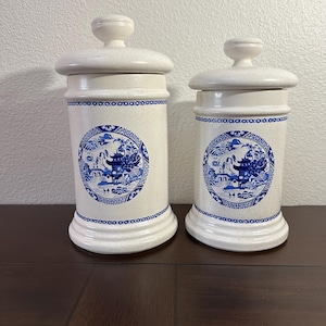 May include: Two white ceramic canisters with blue and white floral designs and lids. The canisters are decorated with a blue and white floral pattern that includes a pagoda, a bridge, and a willow tree.