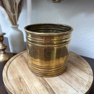 May include: A cylindrical brass planter with horizontal ridges. The planter sits on a round wooden tray. The background includes a white vase with dried flowers, a white candle, and a gold-colored decorative frame.