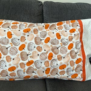 May include: A rectangular pillowcase featuring a repeating pattern of pumpkins in shades of orange, beige, and grey on a white background. The pillowcase has a solid orange band and a white side panel with small grey dots along the right edge.