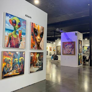 May include: An art gallery featuring framed paintings with alien themes. The paintings depict aliens in various settings, including a beach and a restaurant. The word "Alien" is displayed on the wall. Other artwork is visible in the background.