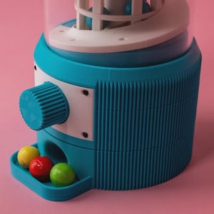 Può includere: Un distributore di palline di gomma turchese e bianco con una parte superiore trasparente, una manopola di erogazione e un vassoio contenente tre palline di gomma gialle, rosse e verdi. La macchina ha una trama a coste e si trova su uno sfondo rosa.
