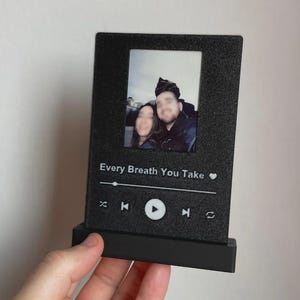 May include: Black photo display featuring a couple's picture. The display includes the text "Every Breath You Take" and a music player design. The display is held by a hand.