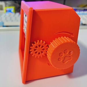 May include: Vibrant orange 3D-printed box featuring a rotating paw print wheel. The box has a rectangular frame and two gears on the side. The wheel has a paw print design. The box is on a white surface.