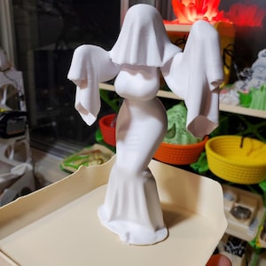 May include: A white, 3D-printed figurine of a draped ghost with outstretched arms. The ghost has a flowing gown and a draped head covering. The figurine is on a beige tray, with other colourful objects in the background.