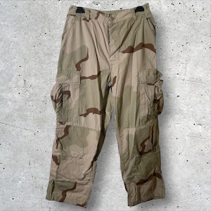 May include: Tan and brown camouflage cargo pants with multiple pockets. The pants have a button and zipper closure and belt loops. The camouflage pattern features irregular brown shapes on a tan background. The pants are made of a durable fabric.
