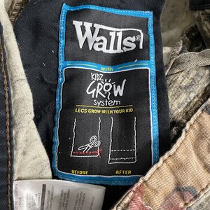 May include: Close-up of a tag on a pair of camouflage pants. The tag reads "Walls" and "Kidz Grow System: Legs Grow With Your Kid." The tag has a blue border and shows a diagram of how to lengthen the pants.
