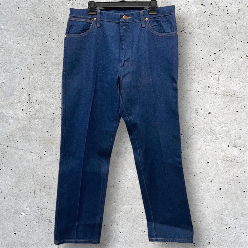 Op de afbeelding: Donkerblauwe denim jeans met een klassiek ontwerp met vijf zakken. De jeans hangen aan een zwarte hanger tegen een gevlekte grijze achtergrond. De jeans hebben een rechte pijp en een knoop- en ritssluiting.