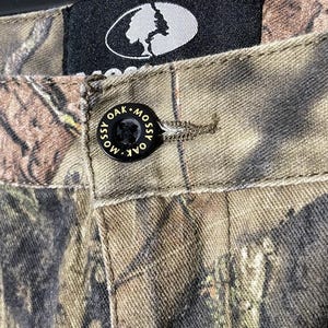 以下が含まれることがあります： 「Mossy Oak」と書かれた黒いボタンが付いた迷彩柄のパンツのクローズアップ。パンツは茶色、緑、タン色の模様です。白い木のロゴが入った黒いタグが見えます。