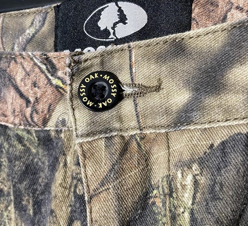 以下が含まれることがあります： 「Mossy Oak」と書かれた黒いボタンが付いた迷彩柄のパンツのクローズアップ。パンツは茶色、緑、タン色の模様です。白い木のロゴが入った黒いタグが見えます。