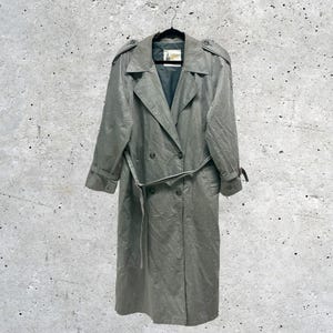 Könnte beinhalten: Ein langer, zweireihiger Trenchcoat in einem gedämpften Salbeigrün. Der Mantel verfügt über einen breiten Kragen, Schulterklappen, lange Ärmel mit Schnallenbündchen und einen passenden Bindegürtel in der Taille. Er hat ein dunkles Innenfutter.