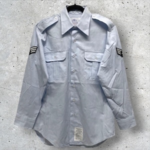 May include: Light blue long-sleeve shirt with two chest pockets and epaulets. The shirt has a button-down front and a collar. Black and white stripes are on the left sleeve. A tag is visible at the bottom of the shirt.