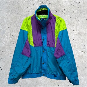 May include: A color-blocked windbreaker jacket featuring a vibrant combination of teal, bright purple, and neon yellow panels. The jacket has a high collar, a front button closure, and long sleeves with elastic cuffs, hanging on a black hanger.