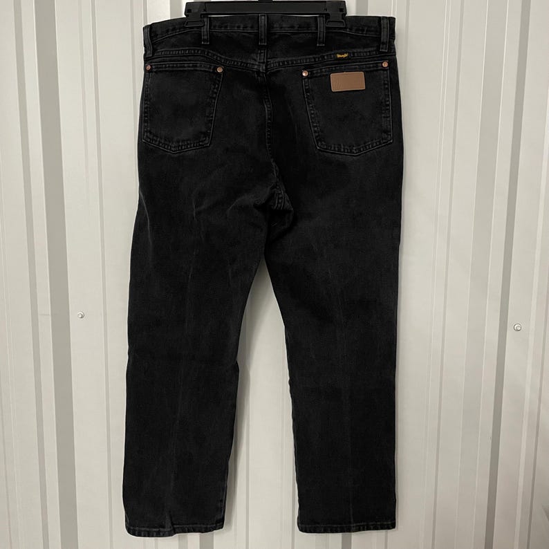 May include: Black denim jeans with a classic five-pocket design. The jeans have a straight leg fit and a brown leather patch on the back. The jeans are hanging on a black hanger.