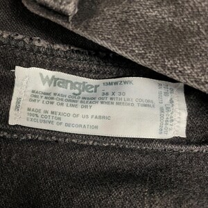 May include: Close-up of a dark gray Wrangler jeans label. The label displays the brand name, size 38 x 30, and care instructions. The fabric is 100% cotton and made in Mexico.