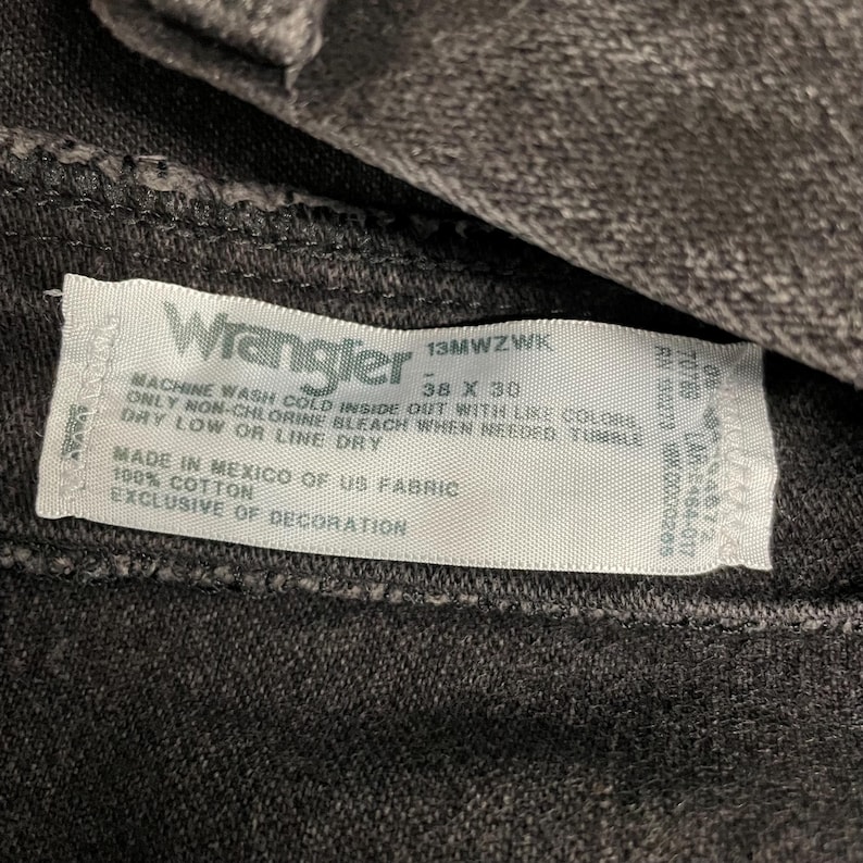 May include: Close-up of a dark gray Wrangler jeans label. The label displays the brand name, size 38 x 30, and care instructions. The fabric is 100% cotton and made in Mexico.