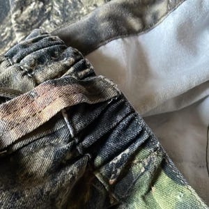以下が含まれることがあります： 緑、茶色、グレーの色合いが混ざった迷彩柄の生地のクローズアップ。伸縮性のあるウエストバンドが見え、詳細なステッチとコントラストのある白い裏地が特徴です。生地はデニムまたは同様の耐久性のある素材のようです。