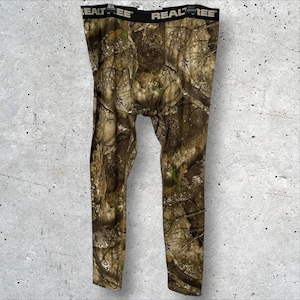 Könnte beinhalten: Tarnhose mit braunem, grünem und beigem Waldmuster. Auf dem schwarzen Bund ist das Wort "Realtree" aufgedruckt. Die Leggings bestehen aus einem dehnbaren Material.