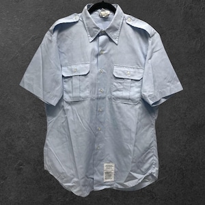 Puede incluir: Camisa azul claro de manga corta con dos bolsillos en el pecho y charreteras. La camisa tiene una botonadura frontal y un cuello. La tela parece ser un material ligero. La camisa está colgada en una percha negra.