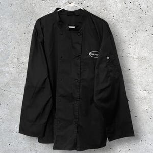 Könnte beinhalten: Schwarze Kochjacke mit langen Ärmeln, Stehkragen und doppelreihigem Knopfverschluss. Die Jacke hat eine kleine Tasche am linken Ärmel und ein Logo auf der Brust. Die Jacke hängt an einem weißen Kleiderbügel.