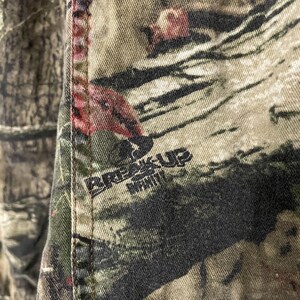 May include: Close-up of camouflage fabric with the words "BREAK-UP INFINITY" printed in black. The fabric features a blend of green, brown, and red hues, with a stitched seam running vertically along the left side.