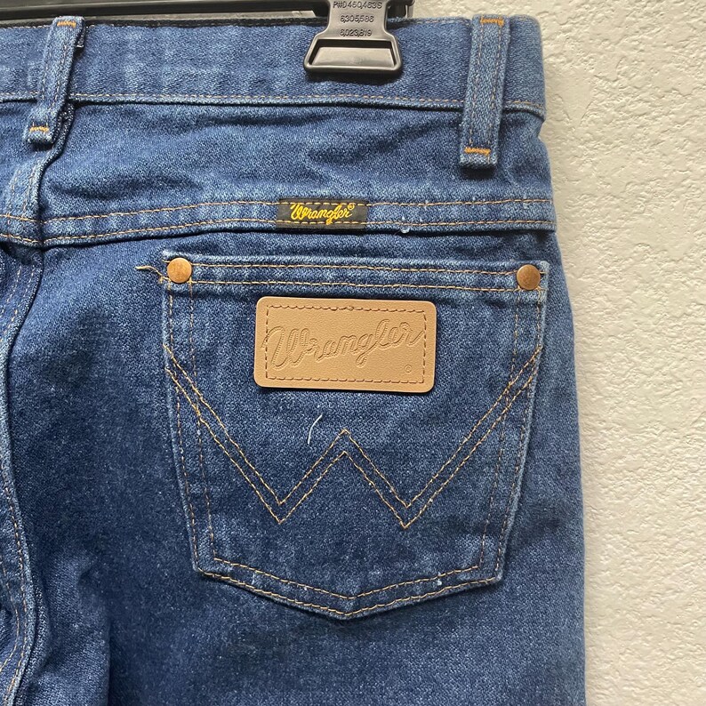 May include: Close-up of blue denim jeans with a Wrangler label on the back pocket. The jeans have a classic five-pocket design with copper-colored rivets and stitching. The Wrangler logo is also visible on the back waistband.