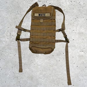 May include: A tan tactical hydration backpack featuring multiple horizontal webbing straps for modular attachments. The top flap displays a rectangular patch with the text WHITE in black lettering. It includes adjustable shoulder straps with plastic buckles.