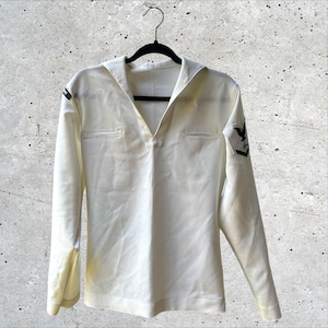 May include: A long-sleeved white sailor shirt featuring a traditional wide collar, a V-neckline, and a black embroidered eagle and chevron patch on the left sleeve. The garment is displayed on a black hanger against a textured gray background.