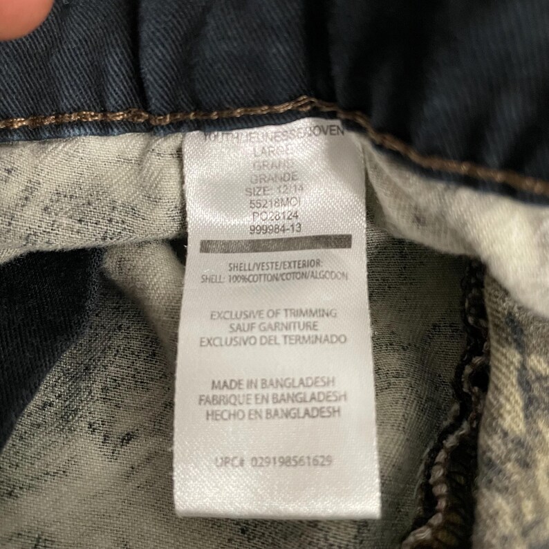 May include: Close-up of a clothing tag with text in English and Spanish. The tag reads "OUTHJEUNESSEJOVEN LARGE GRAND GRANDE SIZE: 12/14" and includes details about the shell material and manufacturing location in Bangladesh.