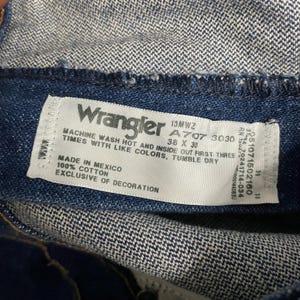 May include: Close-up of a white tag from Wrangler jeans. The tag includes the brand name, size 38 x 30, and washing instructions. The tag is sewn into the dark blue denim fabric. Made in Mexico.