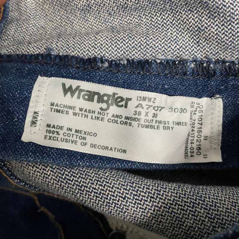 May include: Close-up of a white tag from Wrangler jeans. The tag includes the brand name, size 38 x 30, and washing instructions. The tag is sewn into the dark blue denim fabric. Made in Mexico.