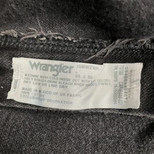 May include: Close-up of a black Wrangler jeans label. The label includes the brand name, size 38 x 30, and care instructions. Made of 100% cotton in Mexico.