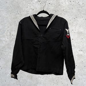 May include: A black long-sleeve sailor shirt featuring a traditional wide collar with three white stripes, buttoned cuffs with white stripes, and an embroidered patch on the left sleeve displaying a white eagle above a red chevron and a small symbol.