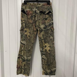 May include: Camouflage cargo pants with multiple pockets and a button and zipper closure. The pants feature a brown, green, and tan woodland camouflage pattern. The pants are hanging on a black hanger against a white background.