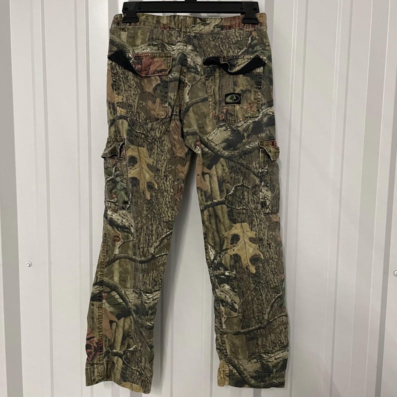 May include: Camouflage cargo pants with multiple pockets and a button and zipper closure. The pants feature a brown, green, and tan woodland camouflage pattern. The pants are hanging on a black hanger against a white background.