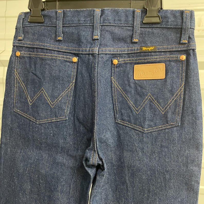Puede incluir: Vaqueros de mezclilla azul oscuro con el logotipo de Wrangler en un parche de cuero. Los vaqueros tienen dos bolsillos traseros con un dise&ntilde;o de costura en forma de W. La costura es de color beige y los vaqueros tienen remaches cobrizos.