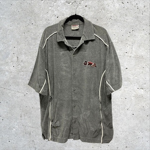 May include: A short-sleeved gray button-down shirt featuring a collar, white piping accents along the shoulders and sides, and a small embroidered graphic on the left chest. The shirt includes a front placket with visible buttons and side pockets.