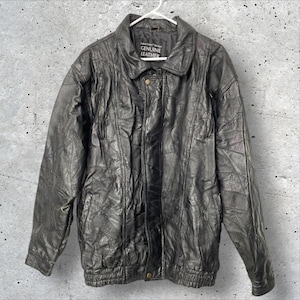May include: Black genuine leather jacket with a collared neckline, button closure, and a ribbed hem. The jacket has a patchwork design and is hanging on a white hanger. The tag reads "GENUINE LEATHER".