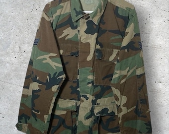 May include: Camouflage pattern jacket with a button-down front, collar, and multiple pockets. The jacket features a mix of green, brown, and black shapes. The jacket is hanging on a white hanger against a gray background.