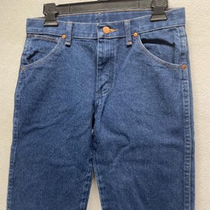 May include: Close-up of a pair of blue denim jeans. The jeans have a classic five-pocket design with copper-colored rivets and a button closure. The denim fabric is a medium wash, and the jeans are hanging on a black hanger.