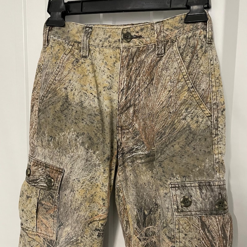 May include: Camouflage cargo pants with buttoned pockets and a button closure. The pants feature a pattern of brown, tan, and green, resembling a forest or woodland environment. The pants are hanging on a black hanger.
