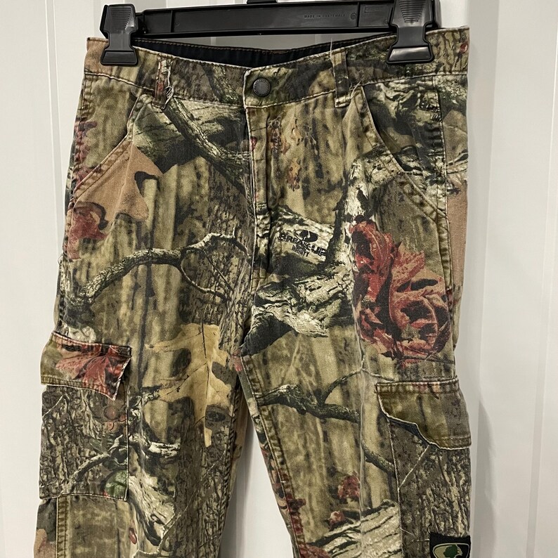 May include: Camouflage cargo pants with multiple pockets, featuring a pattern of brown, green, and tan hues. The pants are hanging on a black hanger. The fabric has a detailed print of branches and leaves.