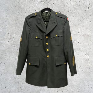 May include: An olive green military-style jacket featuring gold-toned buttons, four front pockets with buttoned flaps, shoulder epaulets, and yellow chevron stripes on both sleeves. A small, colorful embroidered patch is visible on the right shoulder.
