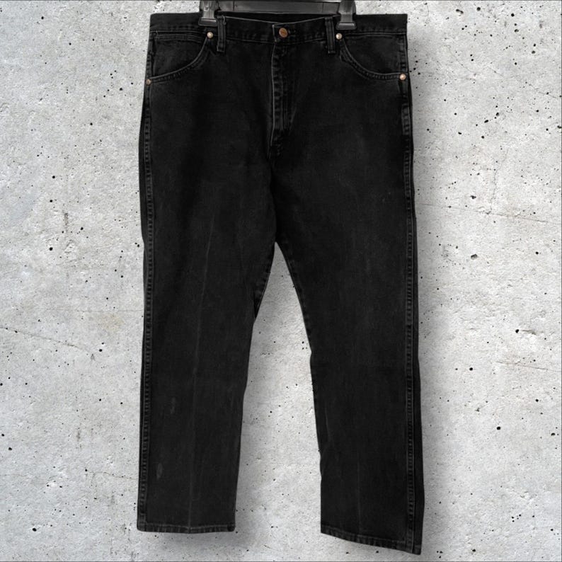 May include: Black denim jeans hanging on a black hanger. The jeans have a classic five-pocket design with a straight leg fit. The denim appears to be slightly faded, giving them a worn look. The jeans are a dark, solid black color.